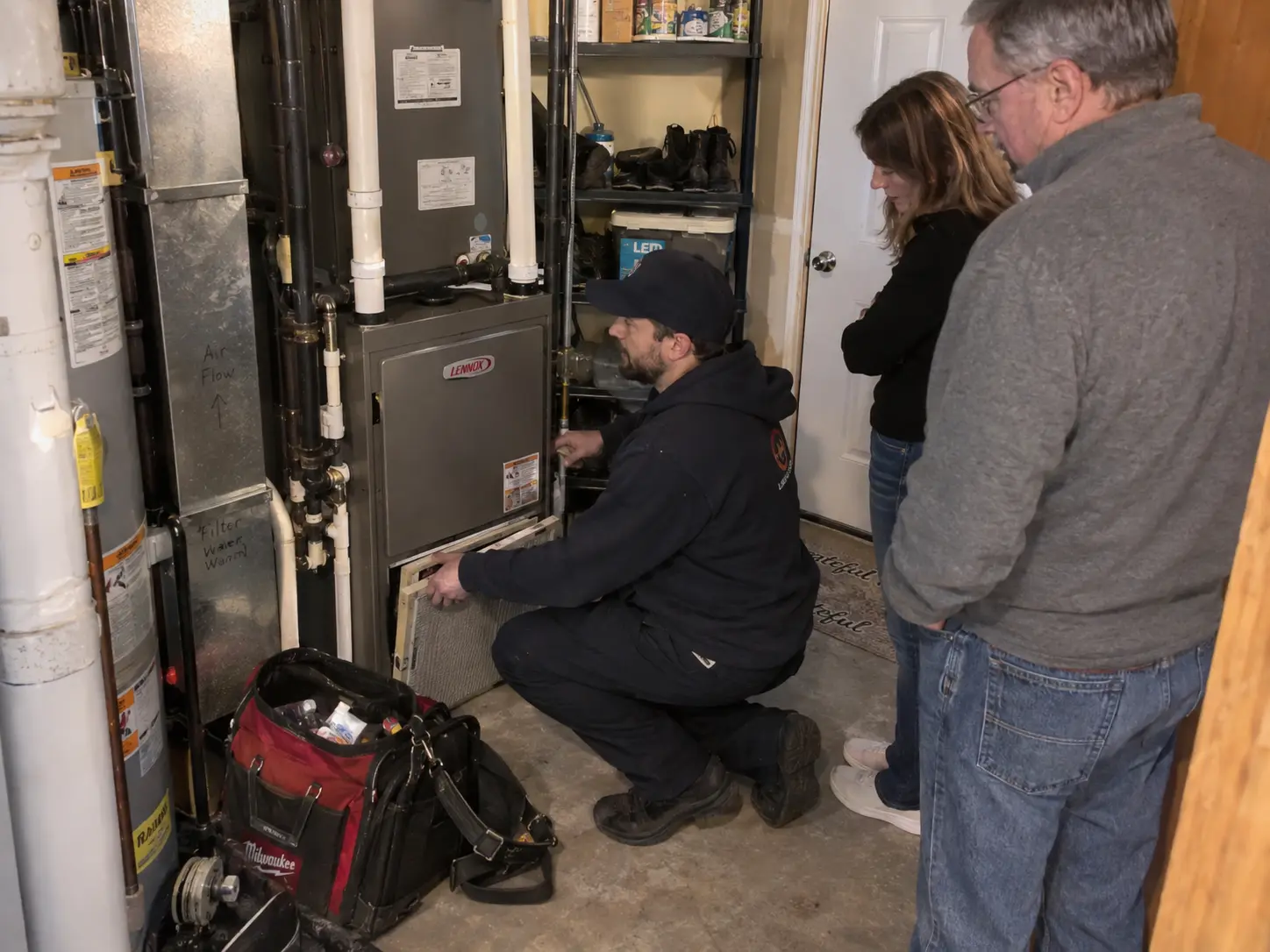 Technician removing a furnace filter while two homeowners look on in a utility area
