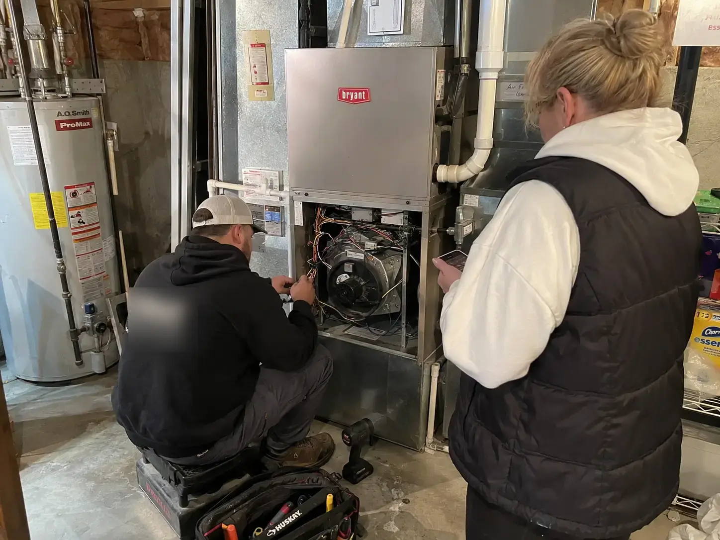 Technician working inside an open furnace cabinet while a homeowner takes notes