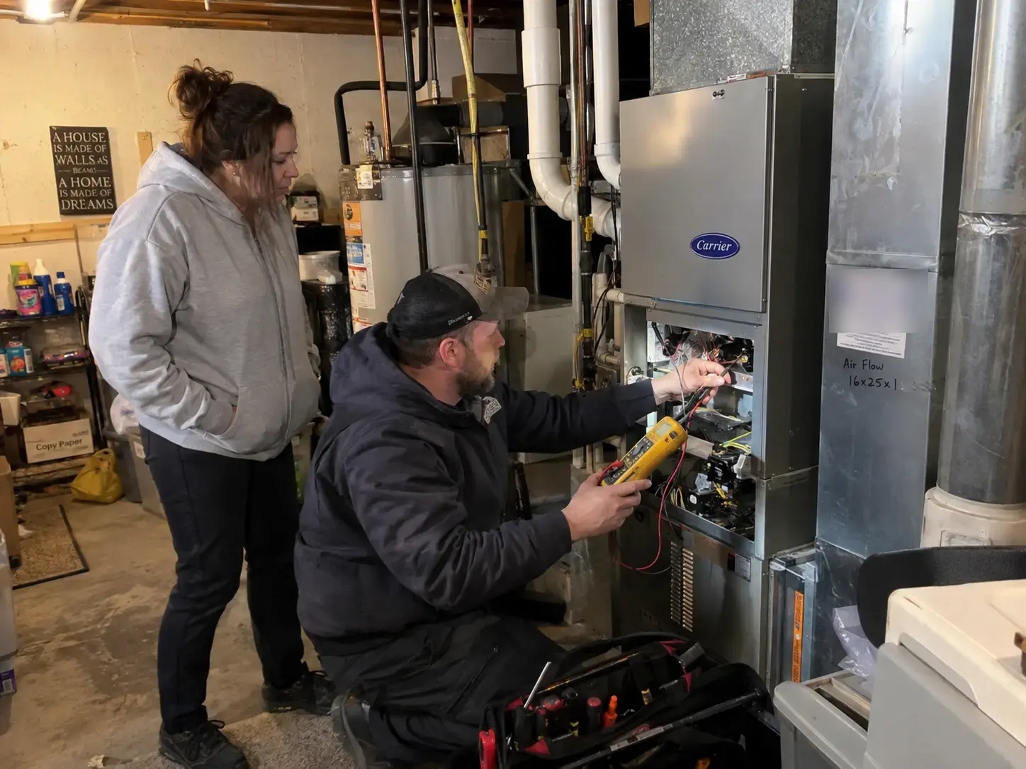 Technician testing furnace wiring with a multimeter while a homeowner watches in a basement